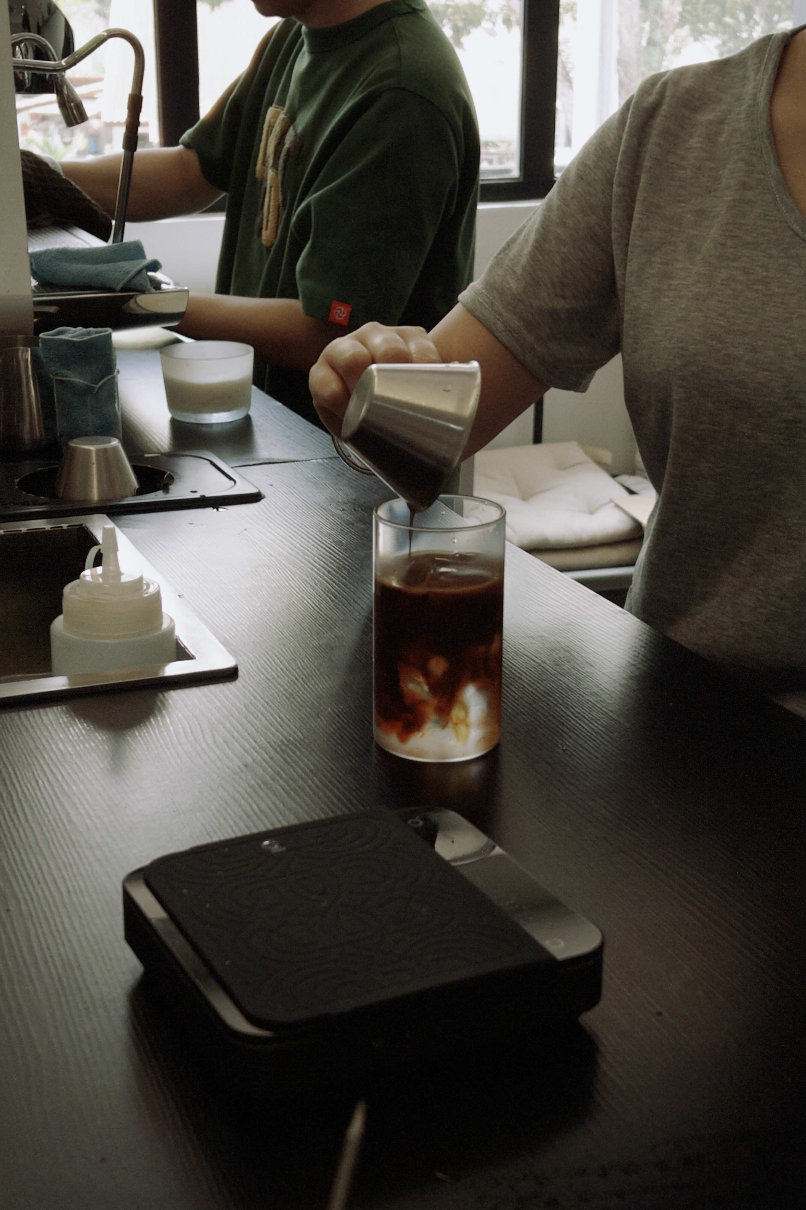 Person pouring a drink at a counter with a glass of iced coffee on the counter.